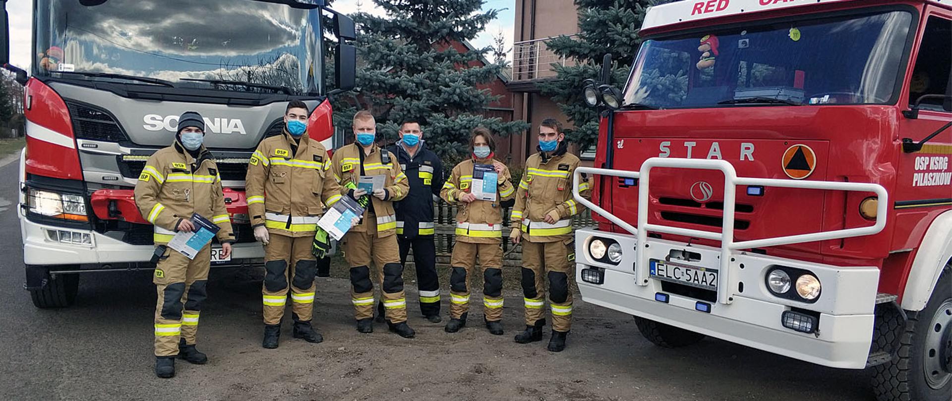 Na zdjęciu widać sześć osób z Ochotniczej Straży Pożarnej ubranych w ubrania typu Moratex barwy jasno brązowej z pasami odblaskowymi koloru szarego oraz jasno-żółtego na tle budynku mieszkalnego. Za nimi rosną trzy drzewa iglaste, po lewej i prawej stronie kadru stoją dwa samochody strażackie barwy czerwonej marki Star i Scania. Ratownicy trzymają w ręce karty informacyjne nt. szczepienia i profilaktyki przeciw COVID-19.