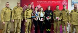 Strażaków oraz pracowników cywilnych stojących i pozujących do zdjęcia. Kobiety trzymają w rękach kwiaty oraz drobny upominek.