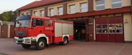 Na pierwszym planie wóz gaśniczy na placu manewrowym przed budynkiem strażnicy. Za samochodem otwarta brama garażowa. Zdjęcie wykonane przed wyjazdem na pożar do Grecji.