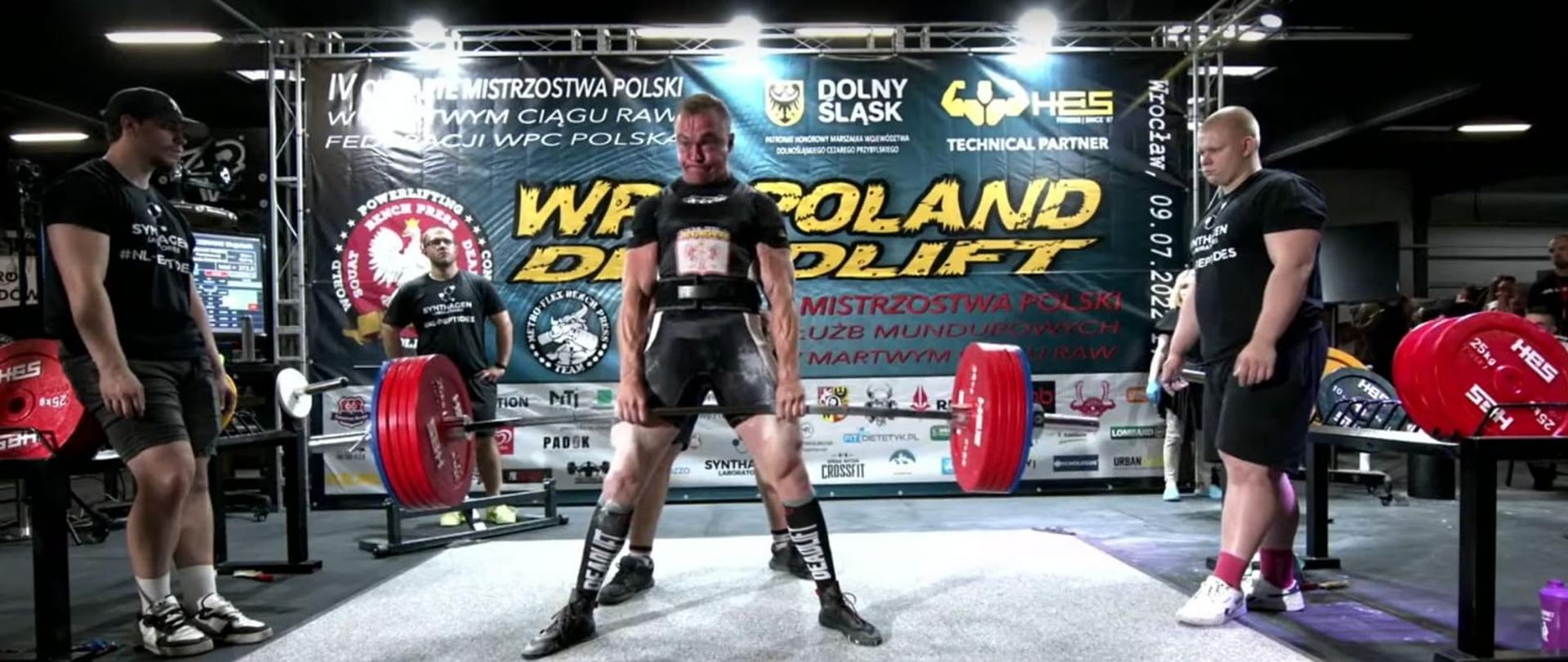 strażak podnosi ciężar w asyście dwóch mężczyzn 