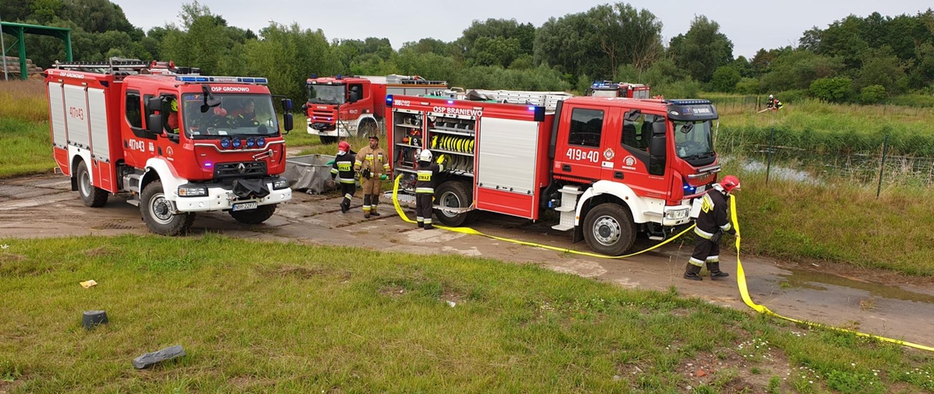 Trzy samochody strażackie ustawiają się przy zbiorniku wodnym, strażacy rozwijają węże.