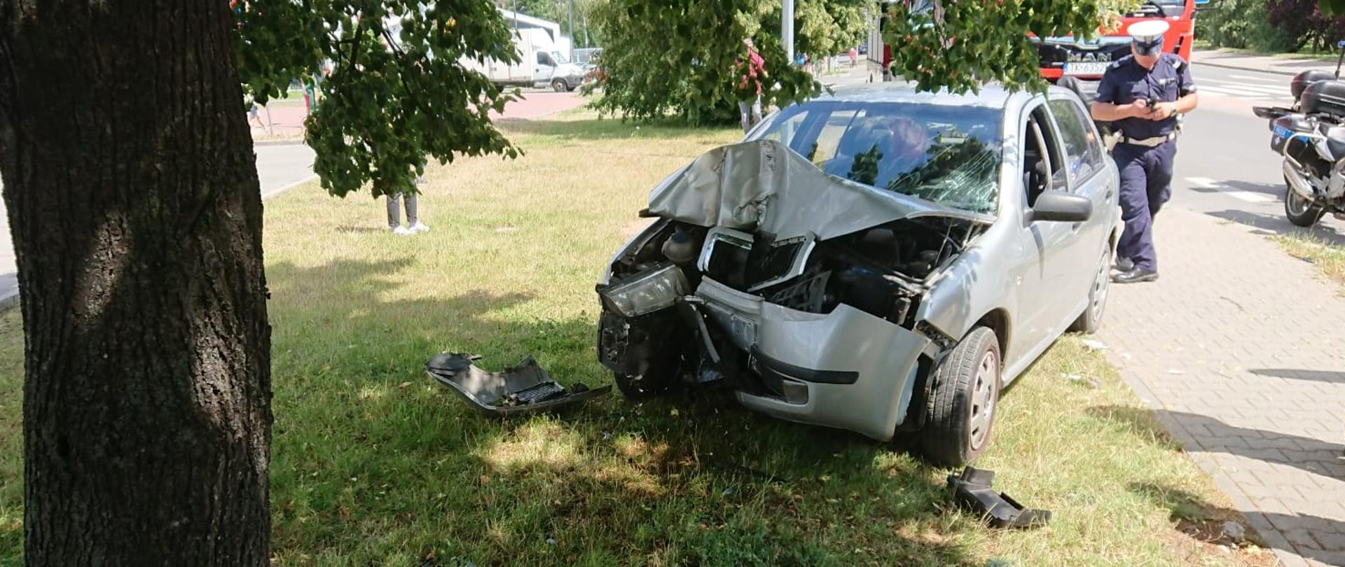 Na zdjęciu widać rozbity przód samochodu osobowego marki Skoda. Samochód stoi przed przed drzewem, w które uderzył nieopodal chodnika. W tle policjant wykonujący czynności dochodzeniowe oraz samochód strażacki. 
