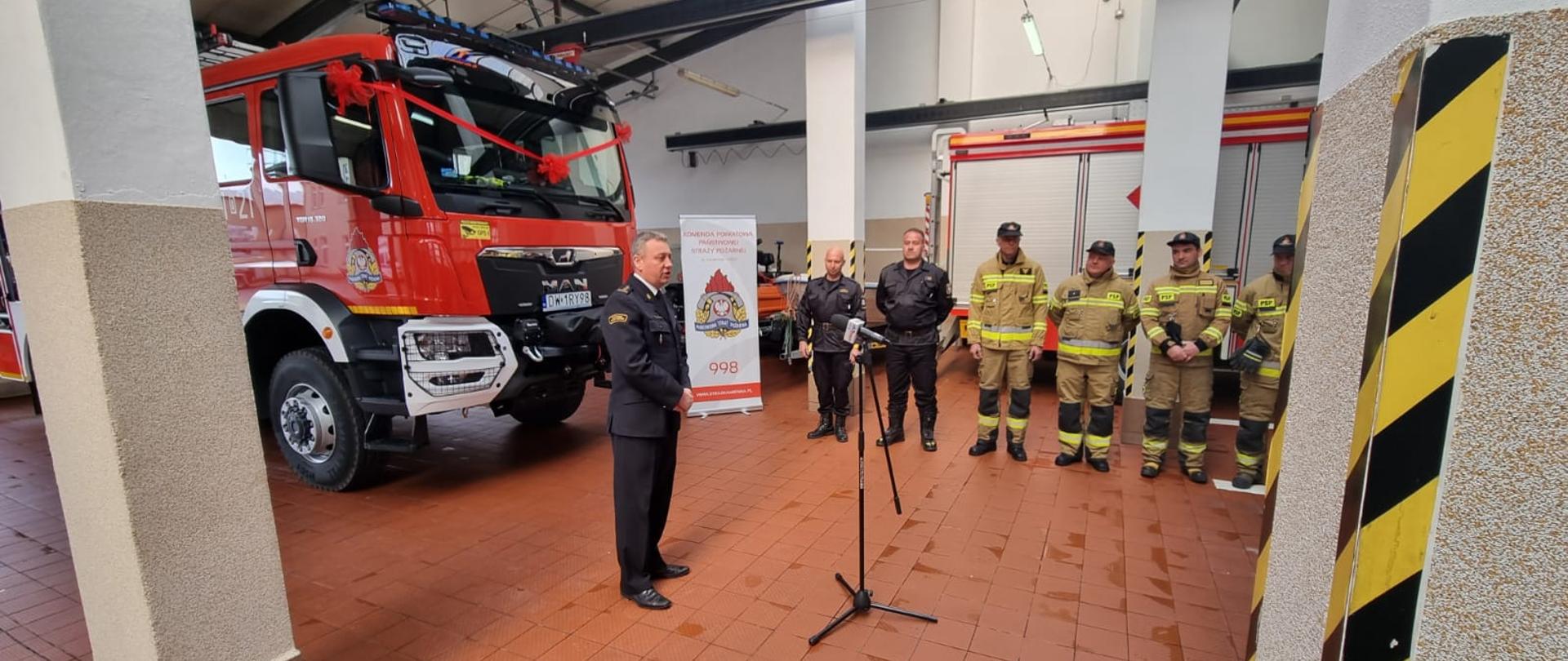 Uroczyste przyjęcie na wyposażenie samochodu ratowniczo gaśniczego w Komendzie Powiatowej Państwowej Straży Pożarnej w Kamiennej Górze. Na planie głównym przemawiający Komendant Powiatowy, za nim widzimy czerwony pojazd ratowniczo gaśniczy. Na drugim planie ustawieni w szeregu w ubraniach specjalnych koloru żółtego.