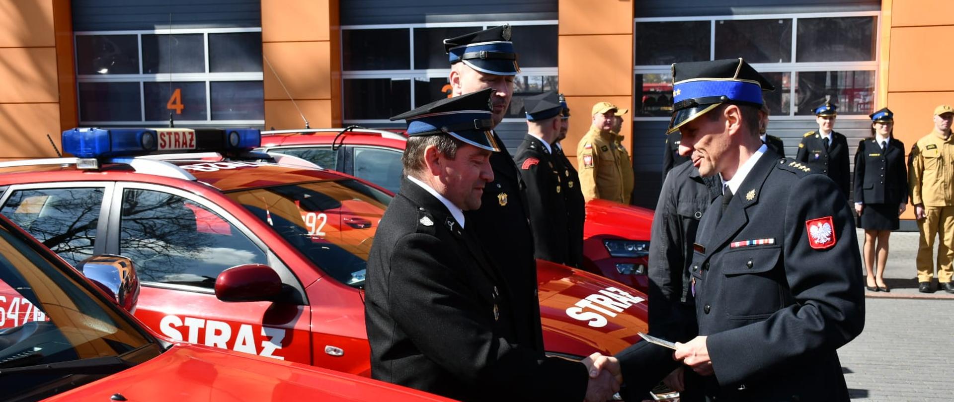 Na pierwszym planie dwóch funkcjonariuszy Państwowej Straży Pożarnej w mundurach galowych podaje sobie ręce podczas przekazania pojazdów. W tle widoczny jest czerwony samochód służbowy z napisem „Straż” oraz budynek jednostki z bramą garażową.