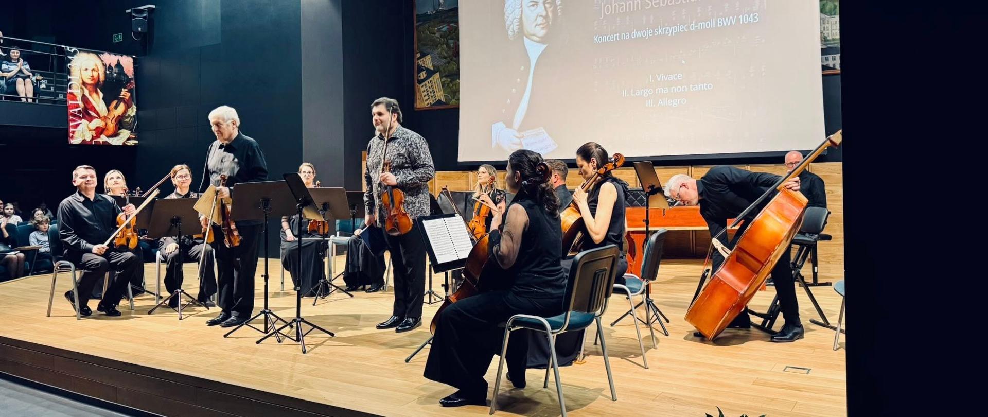 Na zdjęciu widać scenę koncertową, na której występuje zespół muzyków klasycznych. Artyści grają na instrumentach smyczkowych, takich jak skrzypce, altówki, wiolonczele i kontrabas. W tle znajduje się duży ekran z wizerunkiem kompozytora oraz napisem informującym o wykonywanym utworze Johanna Sebastiana Bacha. Muzycy są elegancko ubrani, a publiczność obserwuje występ z widowni. Na pierwszym planie, przy scenie, widoczne są kwiaty, prawdopodobnie przygotowane jako podziękowanie dla wykonawców.