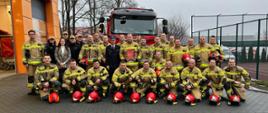 Zdjęcie wykonane na zewnątrz. Przedstawia grupę strażaków w umundurowaniach, stojących na placu przed Komendą. Za nimi auto gaśnicze.