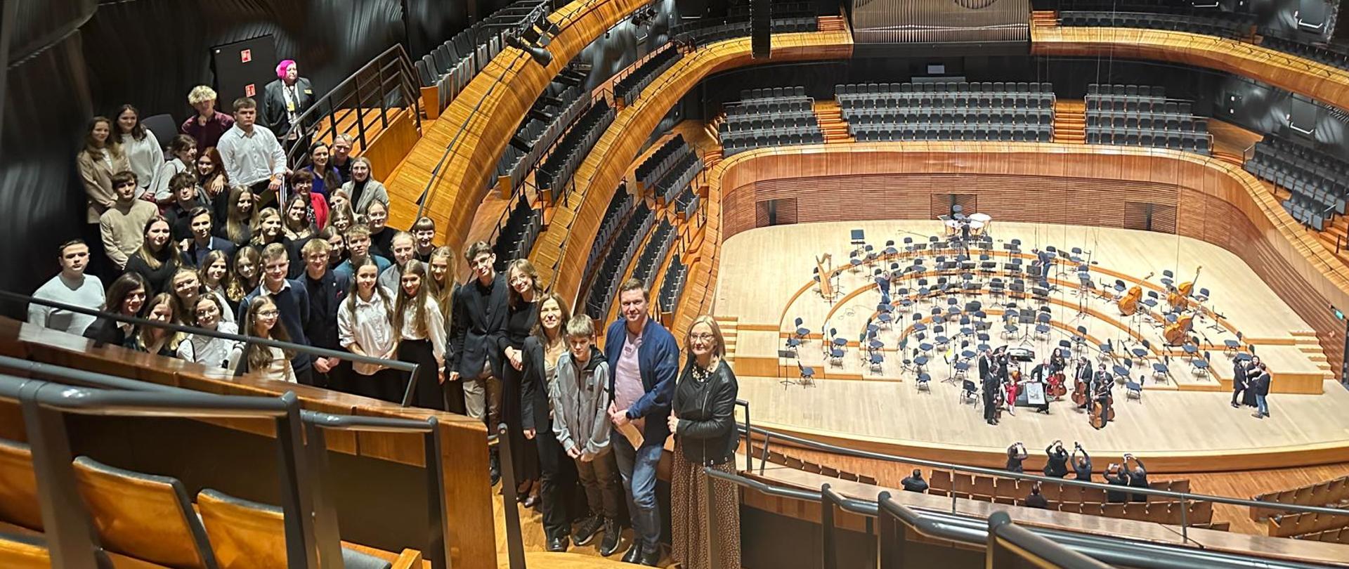 Kolorowe zdjęcie grupowe - Nauczyciele i uczniowie stoją na jednym z balkonów, w tle za ich plecami znajduje się scena NOSPR 