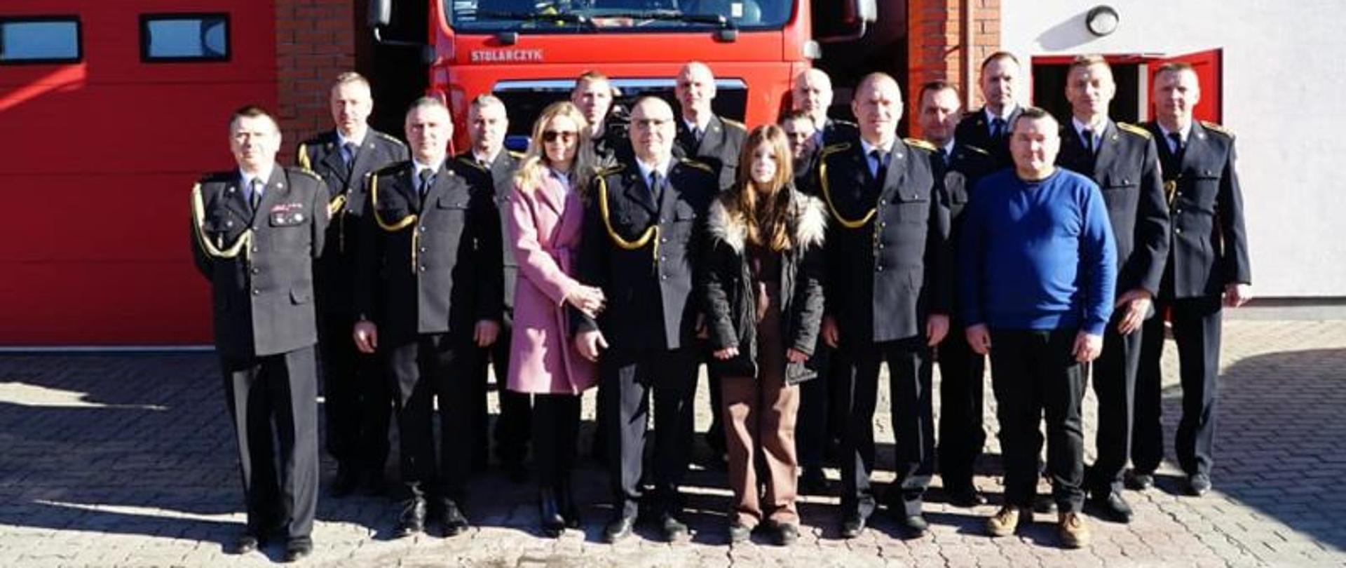 Zdjęcie przedstawia osoby uczestniczące w pożegnaniu odchodzącego na emeryturę strażaka. Wszyscy ubrani galowo, uśmiechnięci. Zdjęcie wykonane na tle garażu oraz pojazdu gaśniczego.