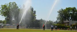 Zawody sportowo-pożarnicze. Płyta boiska na stadionie sportowym. Jedna z MDP w końcowej fazie ćwiczenia bojowego. Rota I i rota II wykonały swoje zadania, a teraz podają w górę strumienie wody. Obok nich członkowie komisji sędziowskiej oceniający konkurencję. W tle drzewa i krzewy.