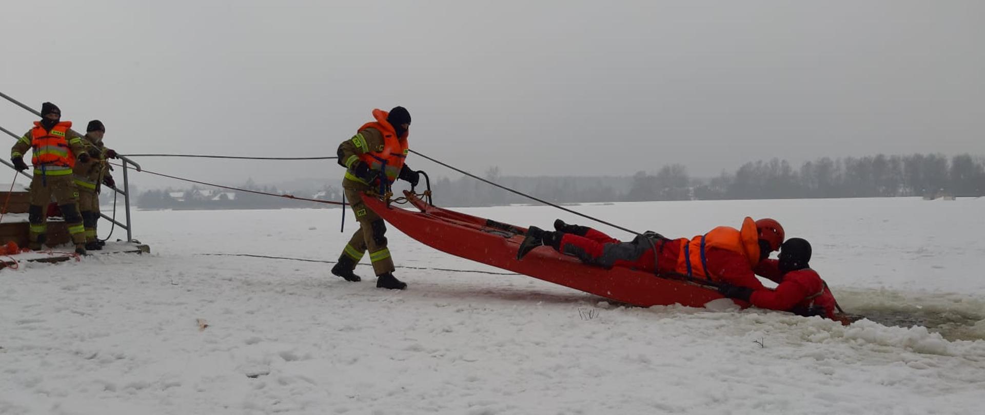 Strażacy w specjalnych skafandrach, zabezpieczeni linkami ratowniczymi ćwiczą podejmowanie poszkodowanego z przerębli z użyciem sań lodowych. Pozoracja realizowana jest na mokro, a strażak znajdujący się w wodzie symuluje poszkodowanego. 