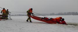 Strażacy w specjalnych skafandrach, zabezpieczeni linkami ratowniczymi ćwiczą podejmowanie poszkodowanego z przerębli z użyciem sań lodowych. Pozoracja realizowana jest na mokro, a strażak znajdujący się w wodzie symuluje poszkodowanego. 