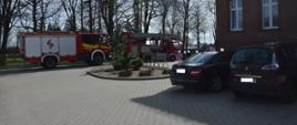 Dziedziniec przedszkola w Bojanowie.W kierunku głównego wejścia do budynku opiekunki prowadzą przedszkolaków, którzy wcześniej zostali ewakuowani na skutek wykrycia zadymmienia. Obok budynku stoją dwa wozy strażackie w tym jeden z podnośnikiem hydraulicznym, a przed obiektem zaparkowane są 2 samochody osobowe.. W tle widać rosnące drzewa.