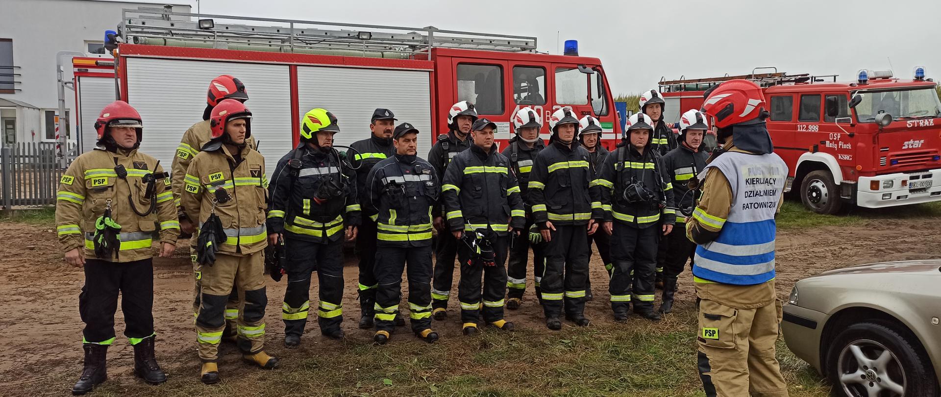 Widać strażaków na tle pojazdów pożarniczych. Strażacy stoją w dwuszeregu, są w pełnym umundurowaniu bojowym.
