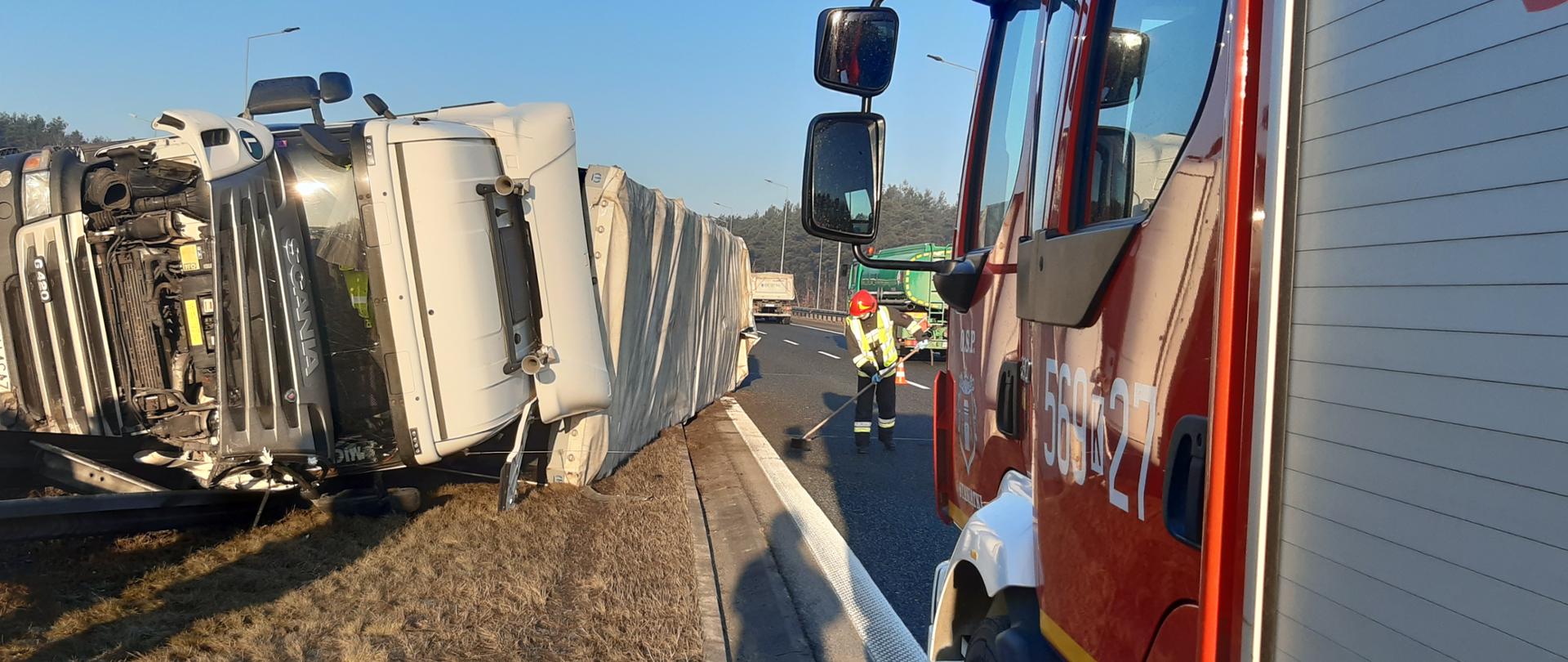 samochód ciężarowy na boku, obok znajduje się samochód ratowniczo - gaśniczy, strażak prowadzący działania na drodze