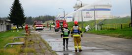 Strażacy ćwiczą gaszenie pożaru zbiornika z paliwem. Widzimy 2 samochody gaśnicze i rozwinięte węże pożarnicze. 1 strażak ubrany jest w ubranie żaroodporne i podaję prąd wody z działka. W oddali widoczne są 2 zbiorniki na o pojemności 10000m3.