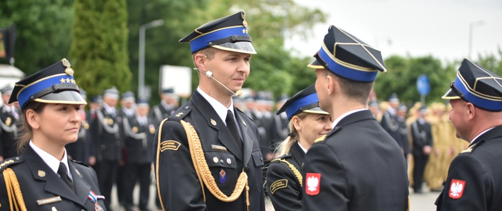 Bliskie ujęcie ceremonii wręczania wyróżnień lub gratulacji. Strażak w galowym mundurze z mikrofonem nagłownym (headsetem) i złotymi sznurami galowymi podaje rękę przełożonemu w czarnych rękawiczkach. Obok stoją dwie kobiety oraz inni strażacy w pełnym umundurowaniu wyjściowym. Na rękawach mundurów widoczne są naszywki z polskim godłem.