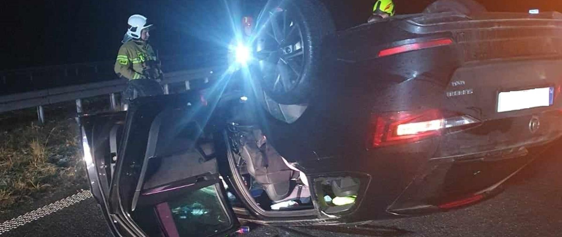 Kolizja na S5. W poprzek pasa ruchu, na dachu leży samochód osobowy koloru czarnego, uczestniczący w kolizji. Drzwi, zarówno od strony kierowcy jak i tylne są otwarte. Na jezdni znajdują sie przedmioty, które wypadły z wnętrza auta (puszka z napojem, chusteczki). Na tle barierek energochłonnych stoi strażak w ubraniu koloru piaskowego i w białym hełmie. Obek niego jest strażak w czerwonym helmie, a zza samochodu widać głowę strażaka w hełmie koloru żółtego. 