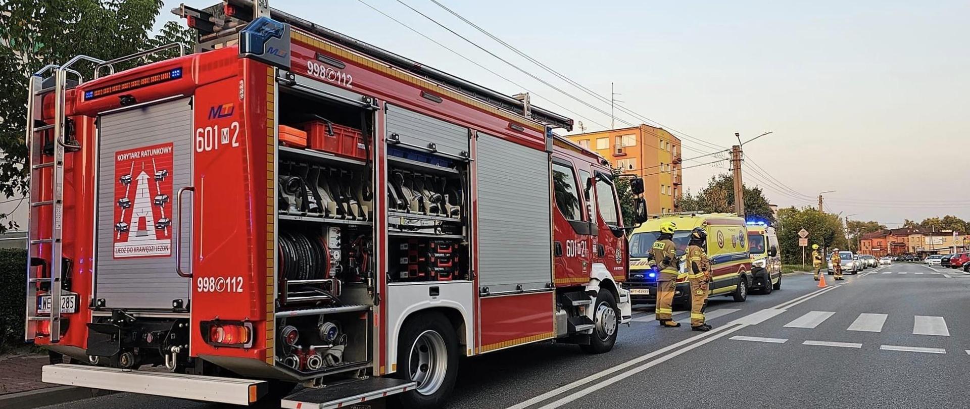 Czerwony wóz strażacki stoi zaparkowany po prawej stronie drogi, z otwartymi bocznymi schowkami, odsłaniając różnorodne narzędzia i sprzęt. Na boku wozu widoczne są napisy „MTI”, „601M21” i „998 112”. Po prawej stronie wozu strażackiego zaparkowane są dwie karetki pogotowia, a obok stoją ratownicy w ubraniach z odblaskami. Droga ciągnie się w tle, gdzie widać więcej pojazdów, a wzdłuż ulicy ciągną się budynki pod bezchmurnym lub częściowo zachmurzonym niebem. Scena sugeruje skoordynowaną akcję ratunkową w warunkach miejskich w okolicy przejścia dla pieszych.