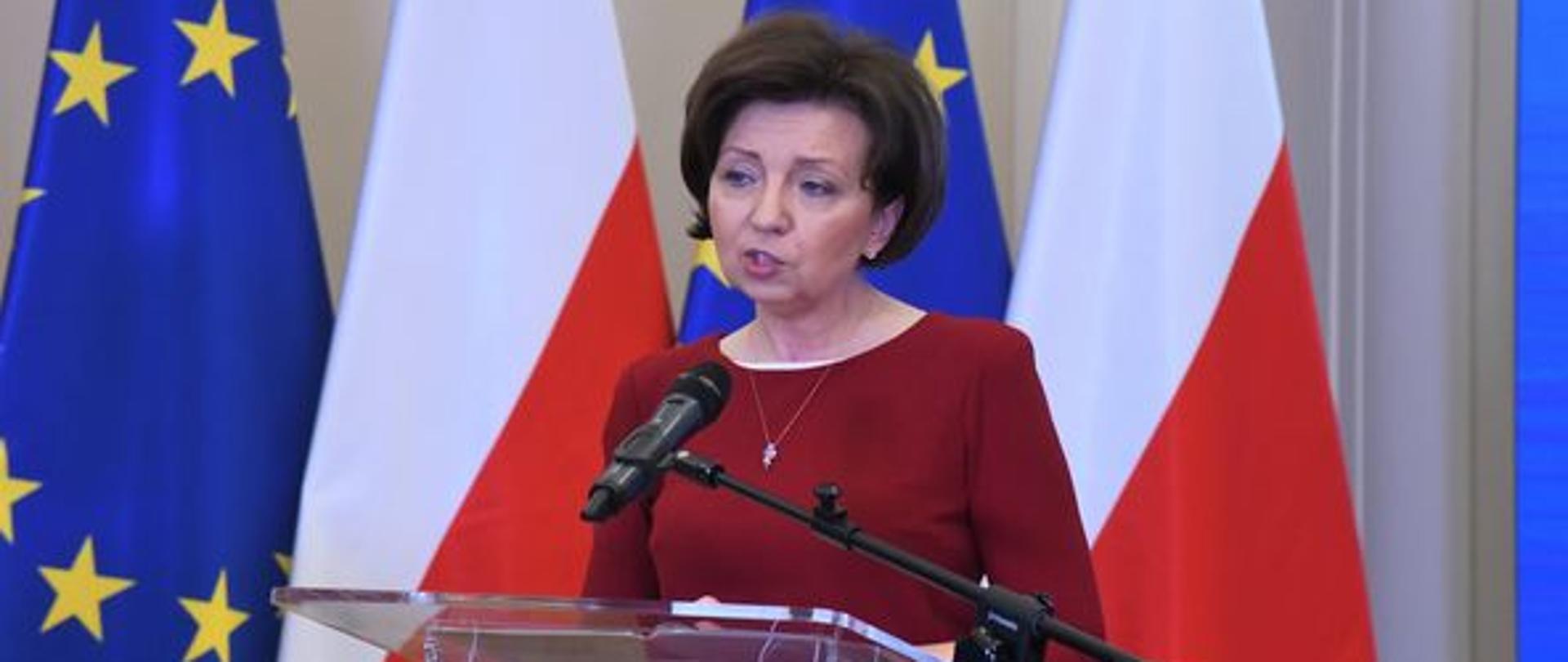 Minister Maląg stoi przy mównicy. Mówi do mikrofonu.