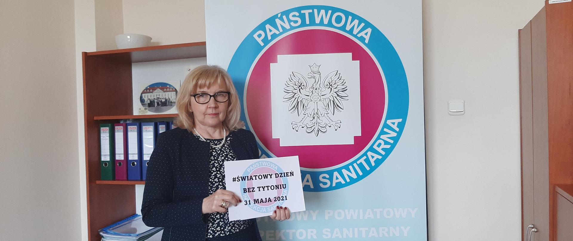 Zdjęcie przedstawia Państwowego Powiatowego Inspektora Sanitarnego w Zgierzu Panią Barbarę Wieteskę na tle logo Państwowej Inspekcji Sanitarnej. W dłoniach trzymającą napis #Światowy Dzień Bez Tytoniu 31 maja 2021. 