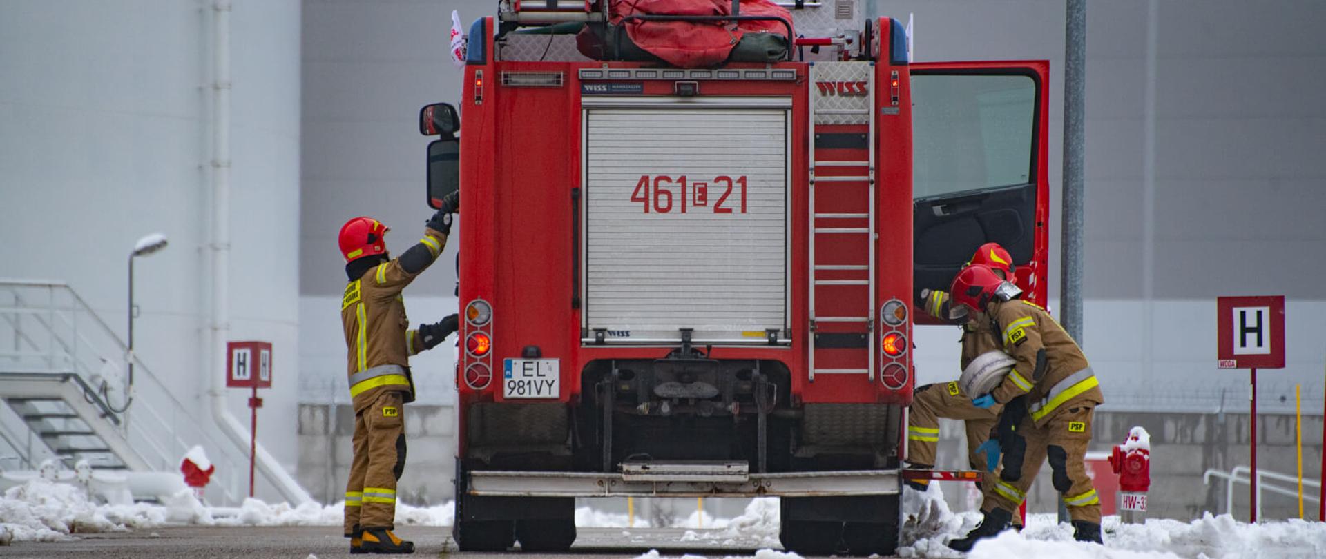 trzech strażaków w ubraniach specjalnych wyciągający sprzęt z samochodu gaśniczego, w tle zbiorniki z paliwem
