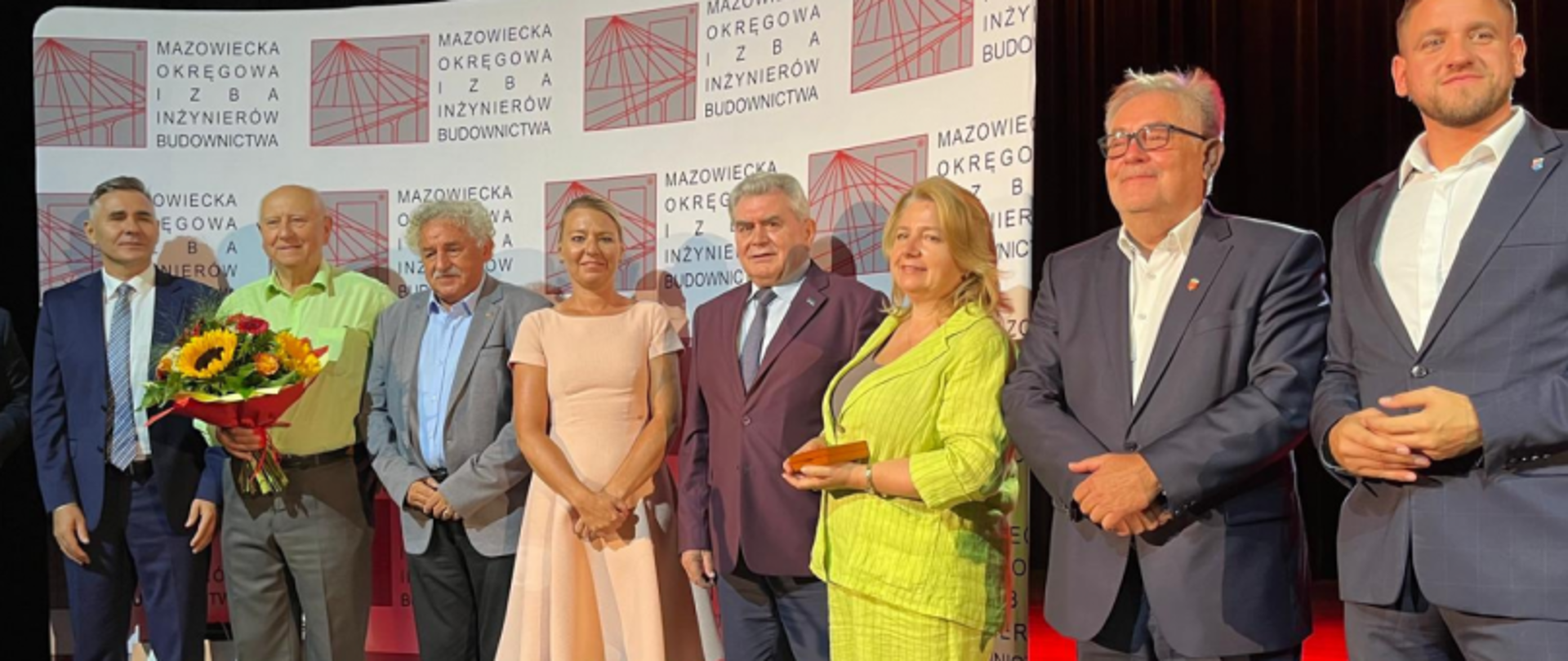 Grupa osób na tle ścianki Mazowieckiej Okręgowej Izby Inżynierów Budownictwa, jedna osoba z bukietem kwiatów.