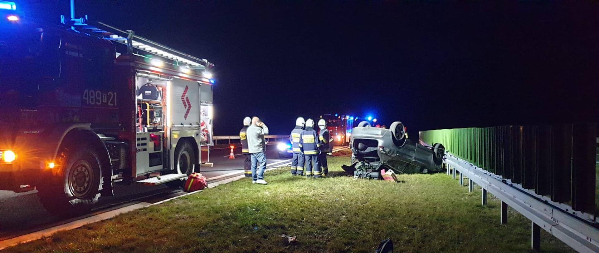 Na zdjęciu widać samochód osobowy który dachował i znajduje się na pasie zieleni na trasie S7. Obok niego stoją strażacy ubrani w ubrania bojowe wraz z poszkodowaną osobą w tym zdarzeniu, która rozmawia przez telefon komórkowy. Obok nich widać samochód strażacki, który jest oświetlony ma włączone sygnały świetlne. W tle widać kolejny samochód strażacki. Zdjęcie wykonano w porze nocnej.
