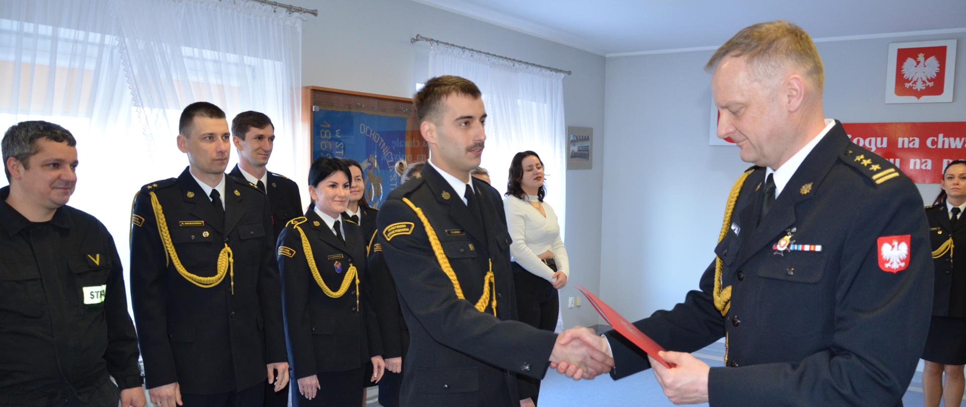Zdjęcie przedstawia moment wręczenia nagrody i gratulacje. W tle strażacy Komendy