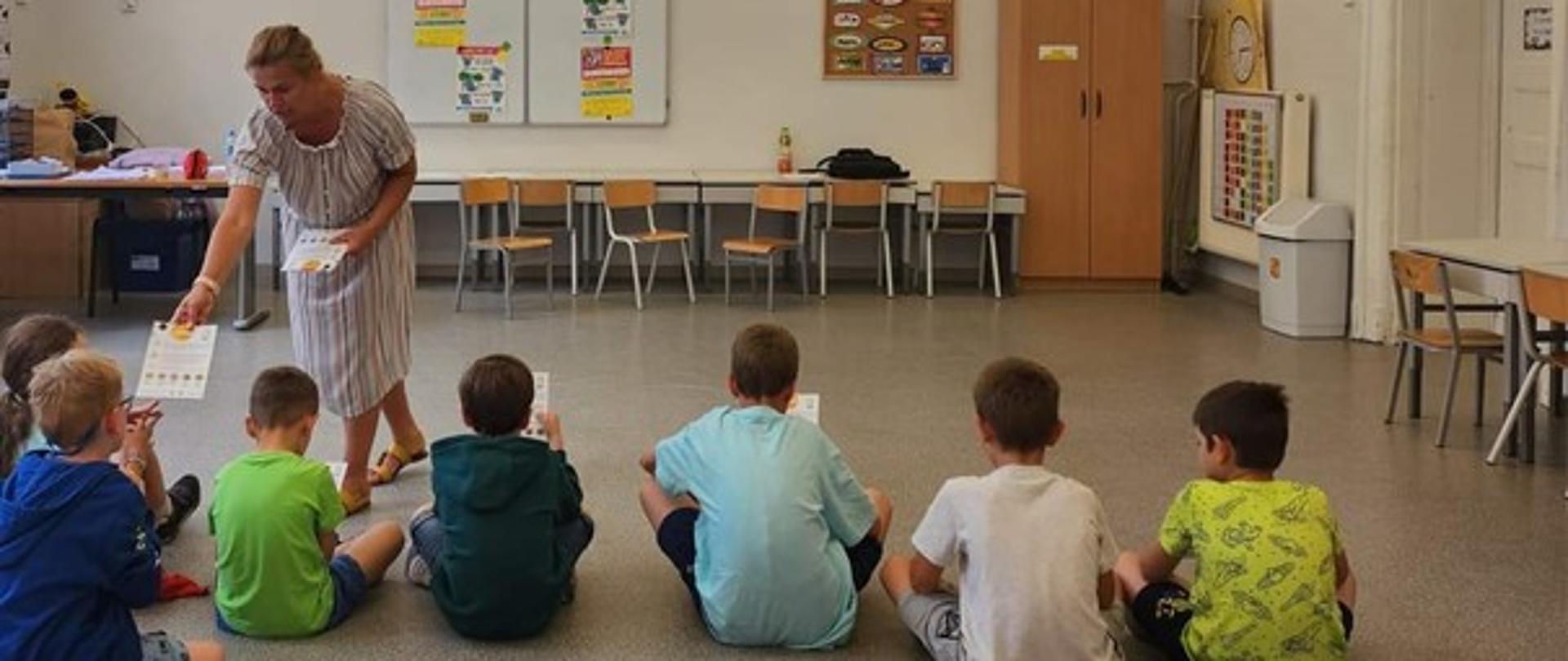 Kobieta w jasnej sukience w kolorowe paski rozdaje ulotki dzieciom siedzącym na podłodze.
