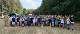 W centrum oraz po prawej stronie kadru stoi stłoczona, liczna grupa kilkudziesięciu obozowiczów w wieku szkolnym. Są ubrani w luźne, letnie stroje sportowe – koszulki, spodenki, czapki z daszkiem i sneakersy. Po lewej stronie, nieco z boku grupy, stoi wyprostowany funkcjonariusz Państwowej Straży Pożarnej w kompletnym, piaskowym (beżowym) umundurowaniu służbowym oraz w czapce z daszkiem.Po prawej stronie, na skraju grupy, stoi starszy chłopak (prawdopodobnie opiekun lub starszy harcerz) w czarnej koszulce z napisem i zielonych szortach, który trzyma w dłoniach kartki papieru i patrzy w stronę grupy. Całe spotkanie odbywa się na suchej, trawiastej polanie. Po prawej stronie grupę otaczają gęste, liściaste drzewa dające cień, natomiast po lewej stronie oraz w głębi widoczny jest las iglasty (sosnowy). W tle, po lewej stronie, można dostrzec elementy bazy obozowej: duży, zielony namiot wojskowy/harcerski, zaparkowany biały samochód dostawczy (bus) oraz inne pojazdy częściowo ukryte między drzewami.