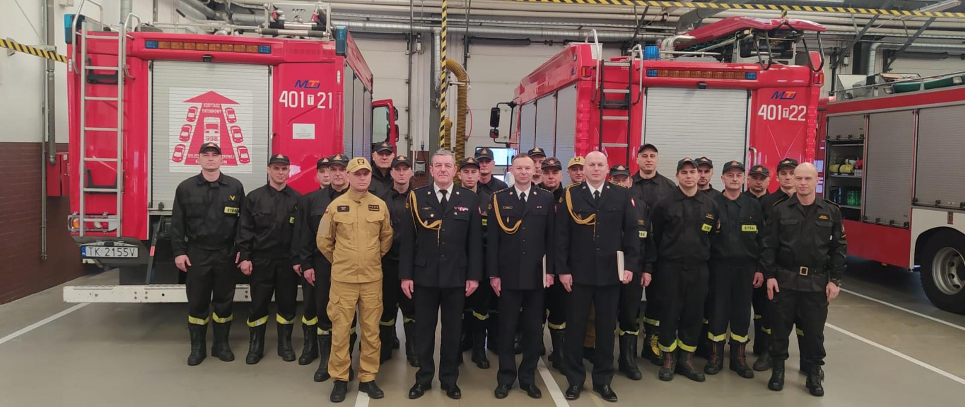 Wspólne zdjęcie strażaków biorących udział w uroczystości mianowania. W tle samochody pożarnicze.