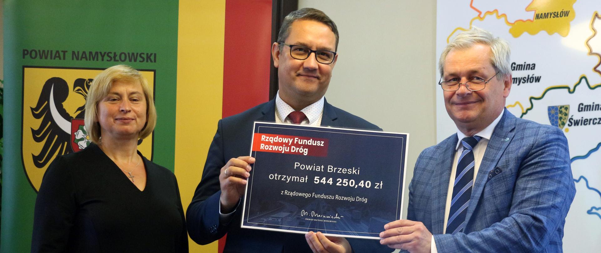 Zdjęcie z uroczystego przekazania czeku samorządom, które skorzystały z Rządowego Funduszu Dróg. Na zdjęciu trzy osoby. Od prawej wojewoda opolski, ubrany w kraciastą marynarkę. Trzyma on wydrukowany czek. Obok niego starosta powiatu brzeskiego, który pozuje do zdjęcia, trzymając fragment czeku. Obok starosty stoi kobieta, ubrana w czarną sukienkę, uśmiecha się. Za tymi osobami, w tle, widać roll up w barwach powiatu namysłowskiego, gdzie odbyła się uroczystość. Za plecami wojewody widać zarys mapy powiatu namysłowskiego.