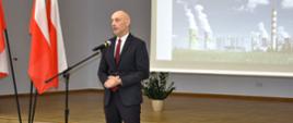 Na centralnym planie widać wojewodę opolskiego, przemawiającego do publiczności. Po lewej stronie kadru biało-czerwone flagi. W tle widać prezentację, wyświetlaną na ekranie. Na niej kominy elektrowni w Opolu. 
