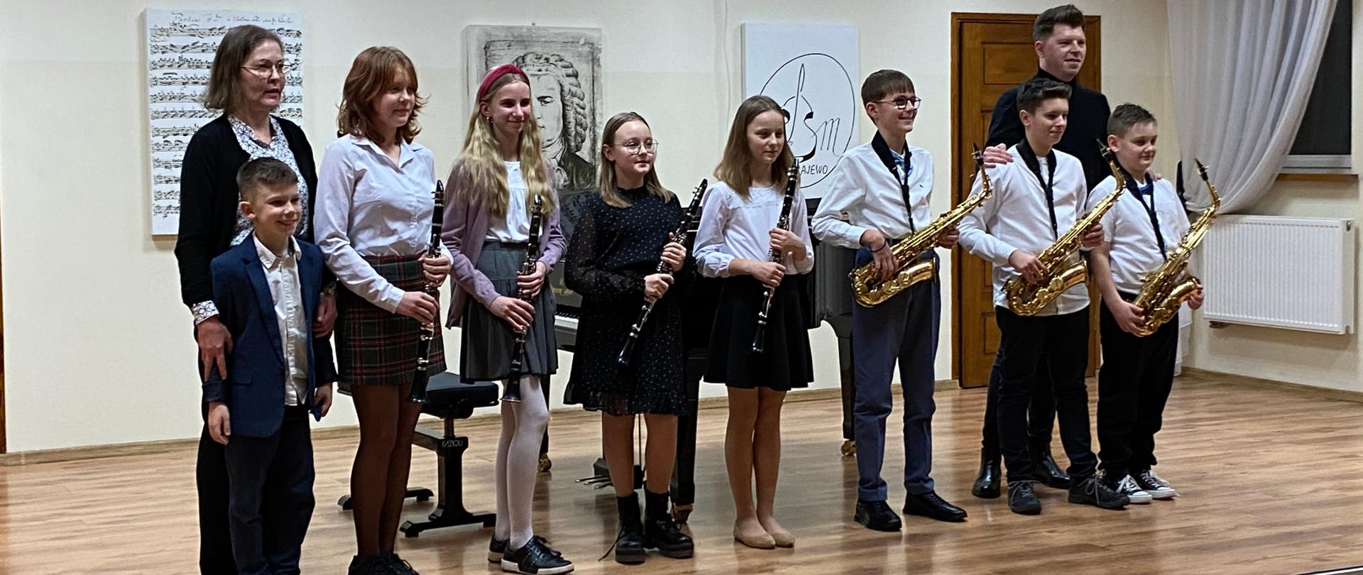 Zdjęcie przedstawia scenę, na scenie ośmioro uczniów z klarnetami i saksofonami. Skrajnie po lewej stoi akompaniator Iwona Czech. Skrajnie po prawej nauczyciel saksofonu i klarnetu Mateusz Kurkowski. W tle na ścianie portret J. S. Bacha oraz obraz z nutami i logiem szkoły. 