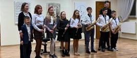 Zdjęcie przedstawia scenę, na scenie ośmioro uczniów z klarnetami i saksofonami. Skrajnie po lewej stoi akompaniator Iwona Czech. Skrajnie po prawej nauczyciel saksofonu i klarnetu Mateusz Kurkowski. W tle na ścianie portret J. S. Bacha oraz obraz z nutami i logiem szkoły. 