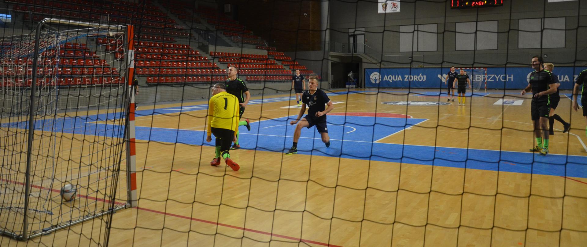 Strażacy ubrani w strojach sportowych w kolorze granatowym oraz czarnym grają w piłkę nożna na hali sportowej