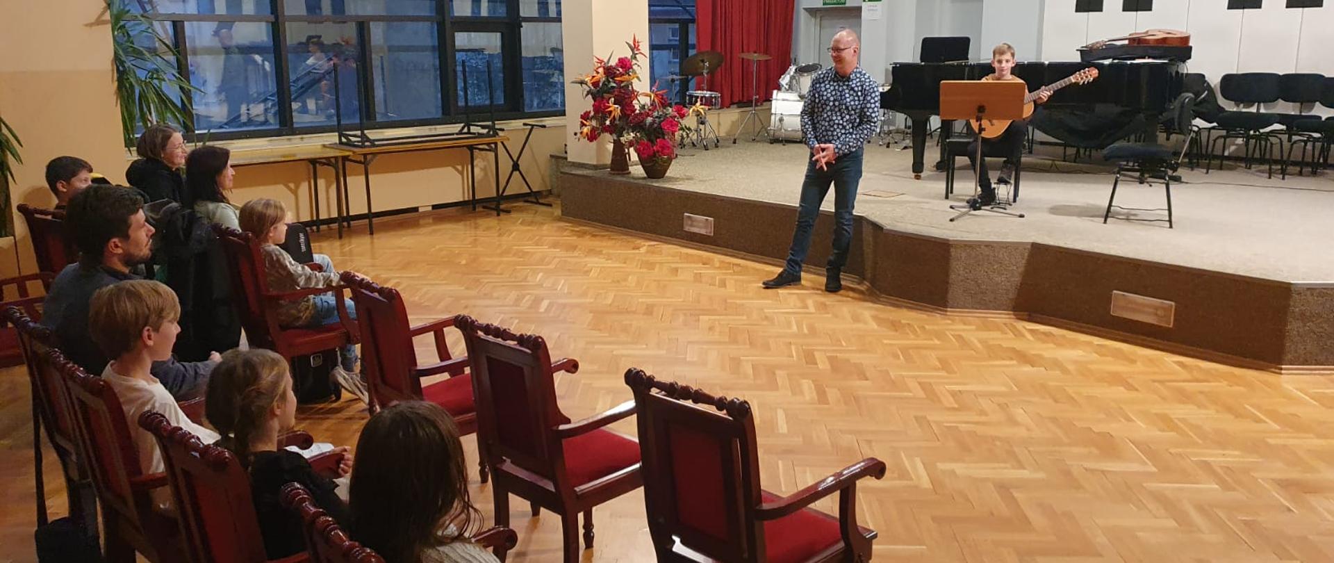 Na podwyższonej estradzie auli PSM za pulpitem siedzi na krześle chłopiec trzymający gitarę, przed estradą stoi mężczyzna i przemawia do publiczności, z przodu widać z boku dwa rzędy widowni z publicznością.