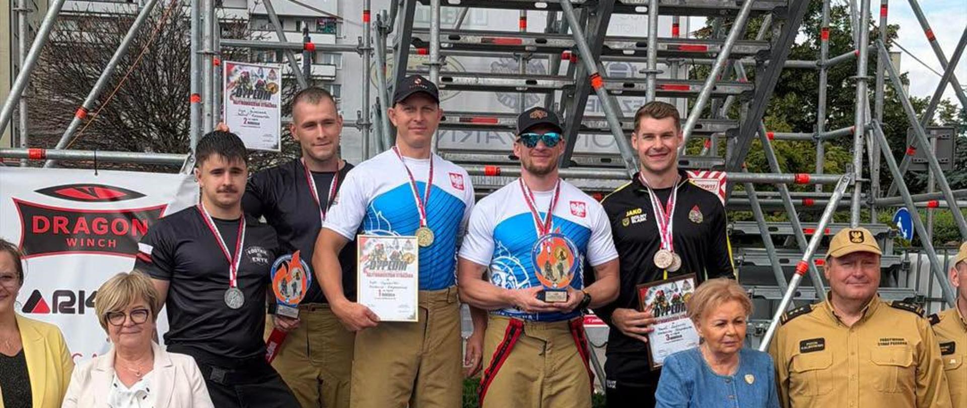 Fotografia przedstawia zwycięzców zawodów pożarniczych, którzy stoją na podium. W tle widać elementy konstrukcji wykorzystanej w zawodach.