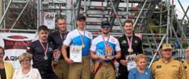 Fotografia przedstawia zwycięzców zawodów pożarniczych, którzy stoją na podium. W tle widać elementy konstrukcji wykorzystanej w zawodach.