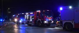 Na zdjęciu widoczne wozy gaśnicze straży pożarnej ustawione wzdłuż ulicy. Samochody mają włączone światła mijania i niebieskie oświetlenie ostrzegawcze. Zdjęcie wykonane w porze nocnej.