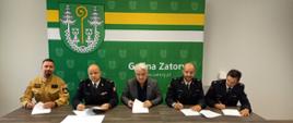 Pięciu mężczyzn siedzi przy stole i podpisuje dokumenty. Dwóch z nich ma mundury strażackie, jeden jasny mundur specjalny, a jeden garnitur. W tle zielona ścianka z herbem i napisem „Gmina Zatory”.