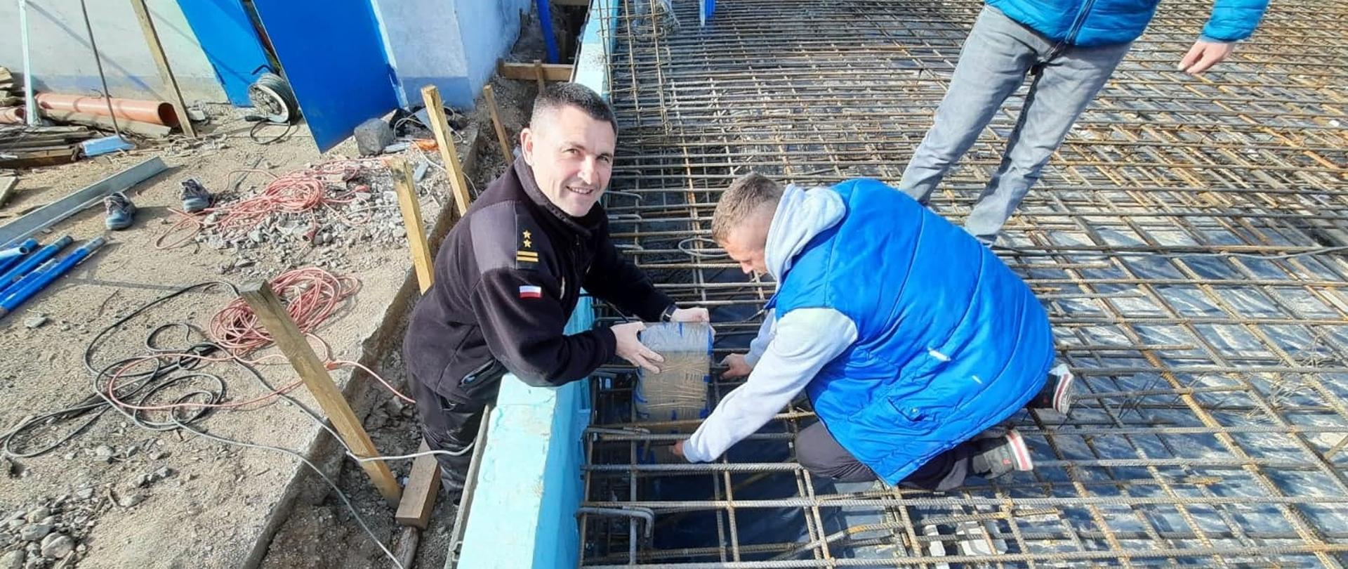 Zdjęcie przedstawia Komendanta Powiatowego Państwowej Straży Pożarnej w Lwówku Śląskim, który wraz z pracownikiem budowy umieszcza kapsułę czasu w fundamencie budynku. 