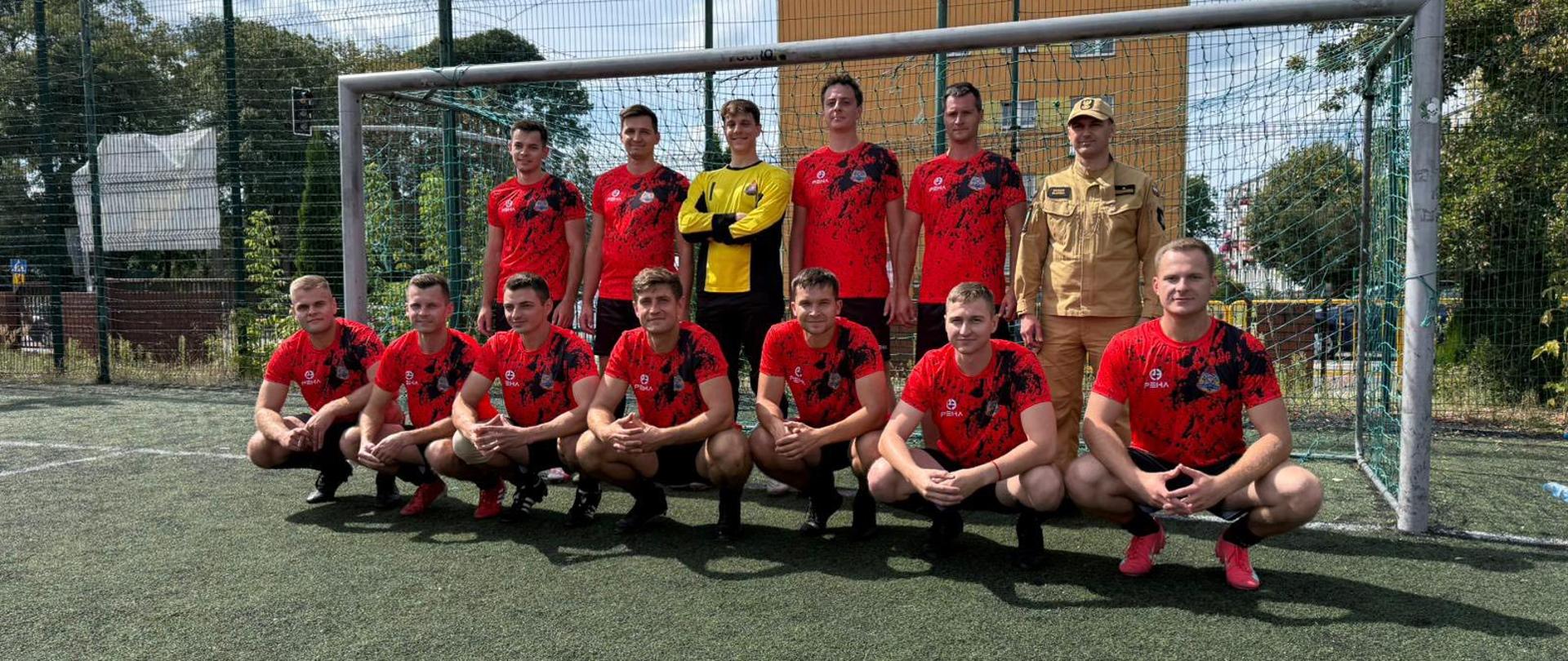 Drużyna piłkarska pozuje do zdjęcia grupowego pod bramką. Część zawodników kuca w pierwszym rzędzie, reszta stoi z tyłu. Wszyscy mają czerwono-czarne stroje sportowe, a bramkarz żółtą koszulkę. Obok drużyny stoi mężczyzna w jasnym mundurze.