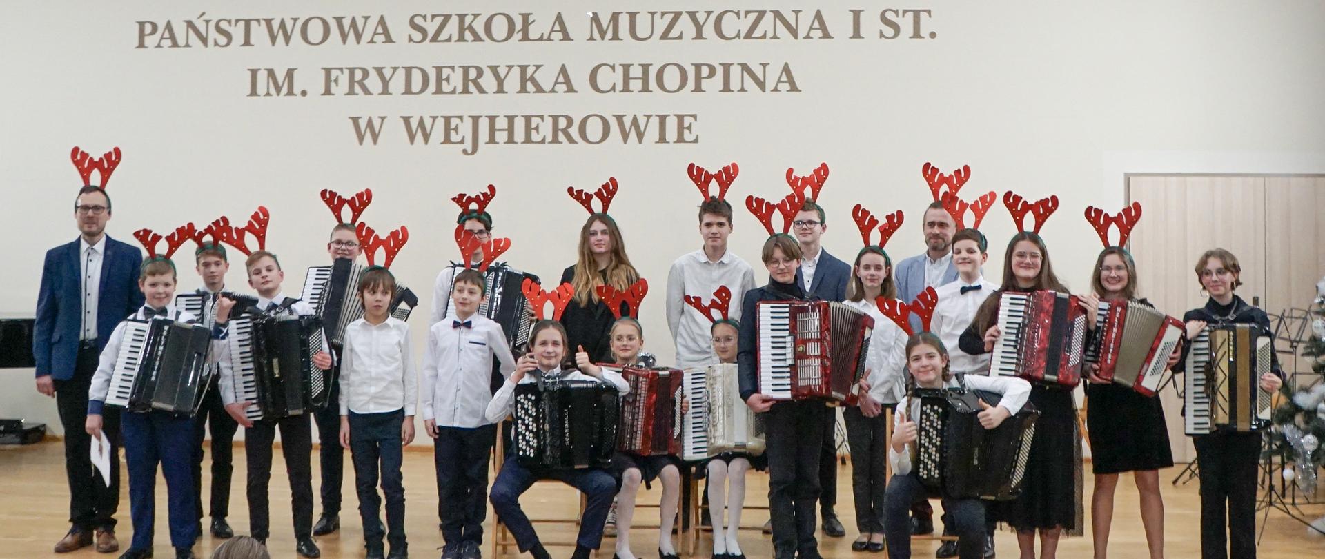 Na zdjęciu widnieją 22 osoby ustawione do zdjęcia grupowego na sali koncertowej. Są to uczniowie oraz dwóch nauczycieli gry na akordeonie. Cześć dzieci pozuje z akordeonami. Nad głowami znajduje się napis "Państwowa Szkoła Muzyczna I st. im. Fryderyka Chopina w Wejherowie". Na dole zdjęcia widnieją dwa rzędy krzeseł oraz część publiczności.