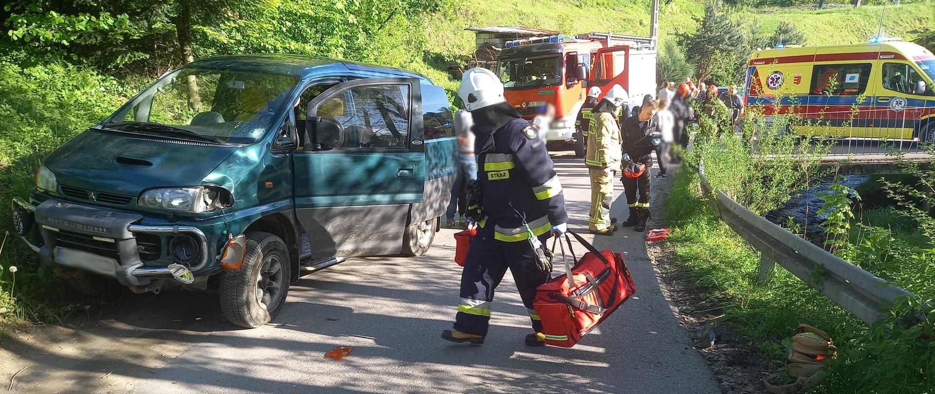 Samochód osobowy, strażak z torbą medyczną idący po drodze gminnej. W tle ludzie na drodze, samochód pożarniczy oraz karetka Pogotowia ratunkowego.