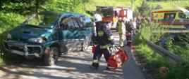 Samochód osobowy, strażak z torbą medyczną idący po drodze gminnej. W tle ludzie na drodze, samochód pożarniczy oraz karetka Pogotowia ratunkowego.