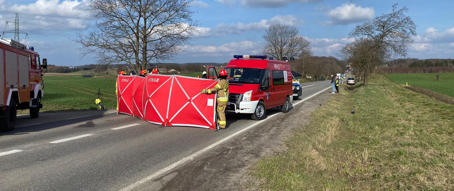 Osłonięte parawanem miejsce wypadku, za którym znajduje się samochód strażacki.