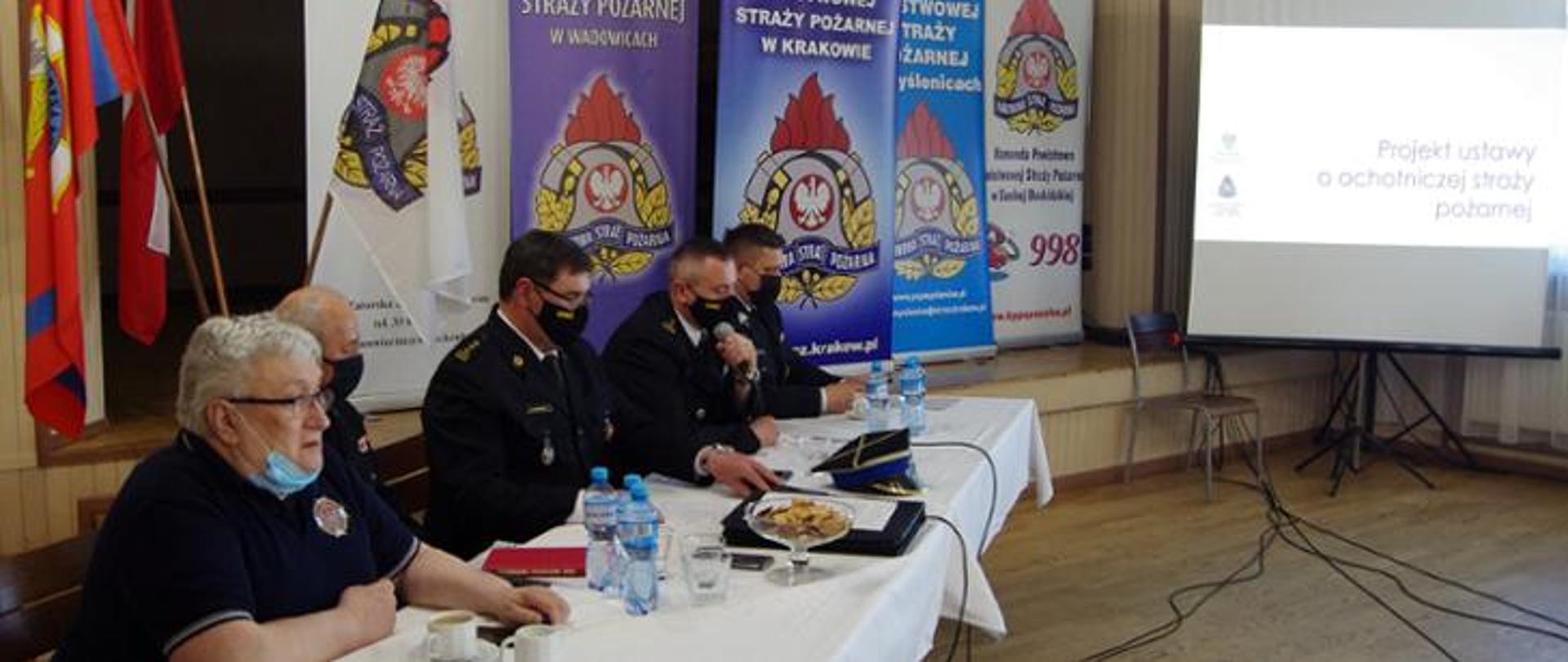 Widok z półprofilu. Na pierwszym widoczny stół prezydialny, za którym siedzą w mundurach strażackich i maseczkach ochronnych oficerowie PSP i członkowie OSP. W centralnej części siedzi Małopolski Komendant Wojewódzki PSP i omawia tekst projektu ustawy o OSP. W tle za stołem prezydialnym widoczne banery KW PSP Kraków i kilku KP PSP. Po prawej stronie ekran z tekstem projektu ustawy o PSP. 