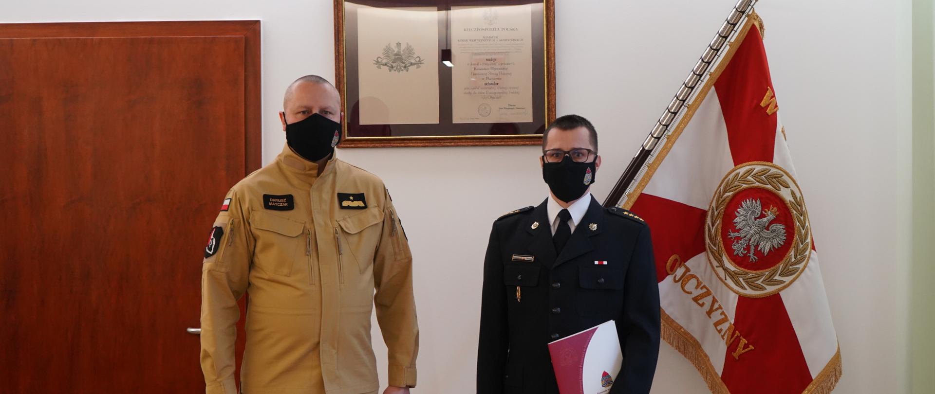 dwóch mężczyzn stoi w maseczkach ochronnych na twarzach. jeden z nich ubrany w miosdowy mundur. drugi w okularach w ciemnym mundurze z białą koszulą , w ręku trzyma teczkę, w tle sztandar