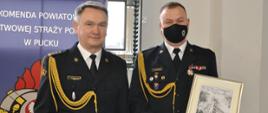 Na zdjęciu stoją dwaj strażacy ubrani w mundury wyjściowe koloru czarnego. PO lewej stronie stoi zastępca pomorskiego komendanta wojewódzkiego PSP w Gdańsku st. bryg. Tomasz Siemiętowski oraz po prawej stronie zdjęcia st. bryg. Andrzej Borkowski, który trzyma obraz - podziękowanie.